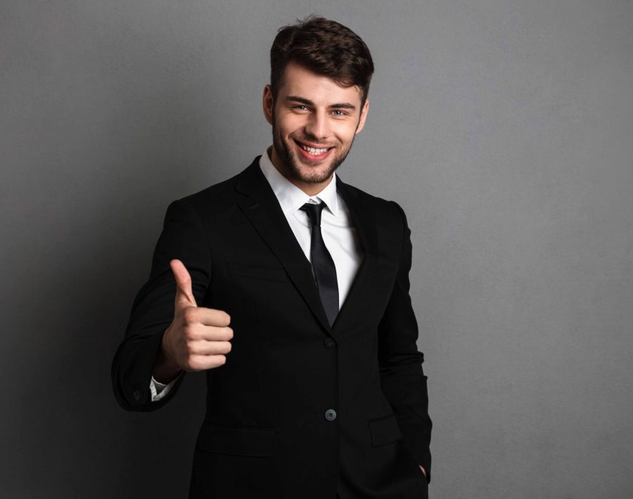 Young successful businessman in formal wear showing thumb up gesture, looking at camera, isolated on gray background